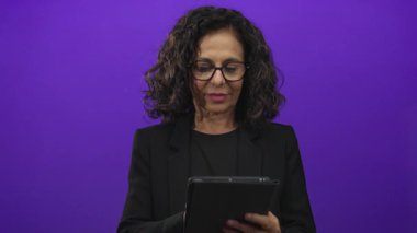 Gözlüklü orta yaşlı kadın, izole edilmiş mor arka planda tablet kullanıyor..