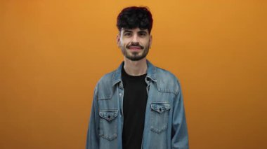 Koyu renk saçlı ve kot ceketli genç adam turuncu bir arkaplanda tuhaf bir ifade sergiliyor..