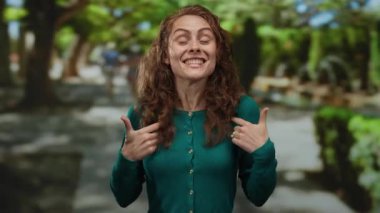 Kıvırcık saçlarıyla parkta gülümseyen, yeşil süveter giyen, açık havada güneşli ağaçların altında mutluluğunu ifade eden bir kadın..