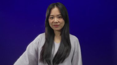 Genç Çinli kadın, kaybedenleri işaret eder. İzole edilmiş mavi arka planda. Sıradan bir süveterle gülümsüyor..