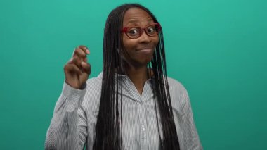 Uzun saçlı ve gözlüklü bir kadın yeşil arka plana karşı jest yapıyor, çizgili gömlek giyiyor, kendine güveni ve mizahı tam..