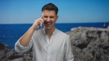 Genç adam deniz kenarında akıllı telefon kullanıyor, güneşli sahil ortamında neşe ifade ediyor..
