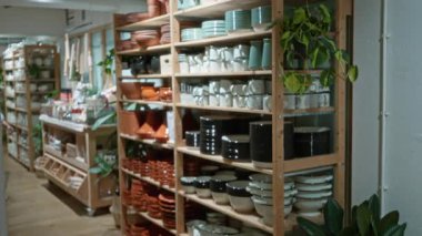 Adam seramik sofra takımlarını ahşap raflarda geziyor. Kapalı bir dükkanda bitki ve doğal ışıkla sıcak ve davetkar bir alışveriş ortamı yaratıyor..