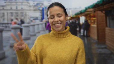 Sarı kazaklı bir kadın, açık hava pazarında ahşap tezgahlar ve hareketli şehir sokaklarında arka planda barış jestleriyle gülümsüyor..