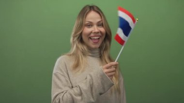 Gülümseyen sarışın kadın, bir fotoğraf stüdyosunda parlak yeşil bir duvara karşı Tayland bayrağı tutuyor..
