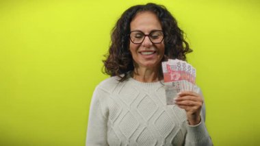 Hong Kong dolarlarını canlı sarı bir arka plana karşı heyecanla tutan kadın gözlük ve rahat bir süveter giyerken neşeli bir ifadeyle para gösteriyor..