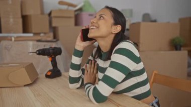 Karton kutularla çevrili telefonda konuşurken gülümseyen İspanyol genç bir kadın ve yeni evinde yeni bir taşınma ya da yenileme olduğunu gösteren bir matkap..