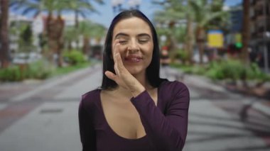 Mor bluzlu genç İspanyol kadın açık havada güneşli bir parkta fısıldıyor. Palmiye ağaçları ve canlı yeşilliklerle çevrili. Rahat bir şehir ortamında neşeyle gülümsüyor..