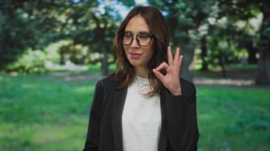 İspanyol kadın iyi bir jest yapıyor ve arka planda ağaçlar olan parka doğru işaret ediyor, gözlük takıyor ve siyah ceket giyiyor, kendine güveni ve pozitifliği yansıtıyor..