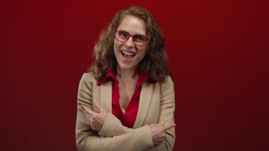 Gözlüklü kadın gülümsüyor ve canlı kırmızı bir duvara başparmak işareti yapıyor. Profesyonel bir ortamda heyecan ve neşe sergiliyor..