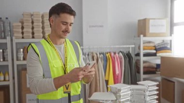 Yansıtıcı yelek giyen genç İspanyol adam bağış merkezinde gönüllü olarak yemek paketleriyle ve elbise raflarıyla çevrili akıllı telefonu kullanıyor..