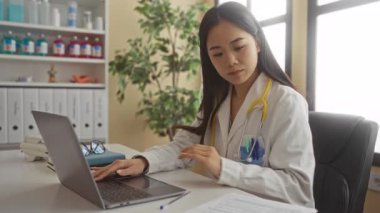 Steteskoplu genç bir kadın doktor dosya ve tıbbi malzemelerle dolu bir klinikte dizüstü bilgisayar üzerinde çalışıyor..
