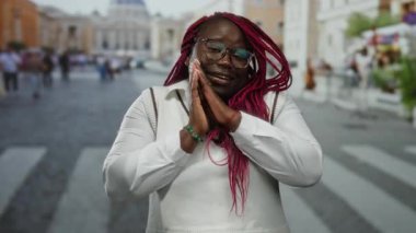 Pembe örgülü gülümseyen kadın, elleri uyku jesti ile Vatikan şehir meydanında poz veriyor, sokak manzarasını ve kültürel çevreyi gösteriyor..