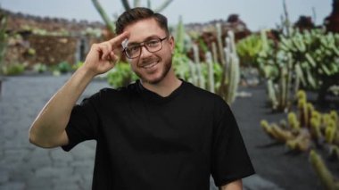 Park ortamında gülümseyen sakallı genç adam, alnında parmakları olan, gözlüklü ve siyah gömlekli, yeşil bitkiler ve taş duvarlarla çevrili..