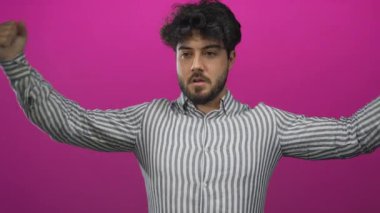 Sakallı ve çizgili gömlekli genç adam canlı pembe arka plana karşı kaslarını esnetiyor..