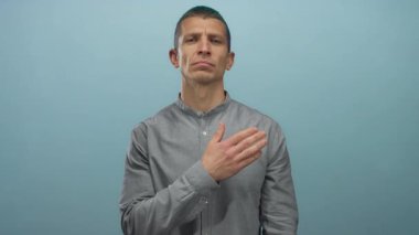 Gri gömlekli adam, mavi duvarlı arka planda yemin jesti yapıyor. İçtenlik ve ciddiyeti ifade ediyor..