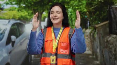 Turuncu yelek giymiş gönüllü bir kadın yeşil ağaçlar, kapalı gözler ve odaklanmayla eski bir kasabanın güneşli sokaklarında sakinliği ve farkındalığı sembolize eden jestler yapıyor..