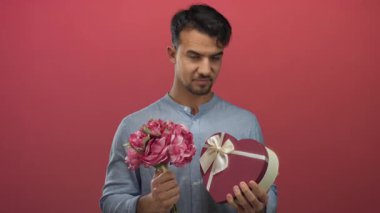 Kırmızı arka plan üzerinde çiçek ve hediye kutusu tutan genç adam romantizm ve düşünceli.