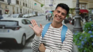 Şehir caddesinde gülümseyen genç adam çizgili tişört giyiyor başparmağıyla cana yakın ve cana yakın görünüyor şehir hayatı tarzını ve açık havada pozitif titreşimleri gösteriyor..