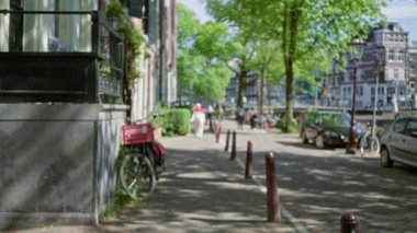 Amsterdam caddesinde yürüyen insanlar, park etmiş bisikletler, ağaçlar, klasik mimari ve güneşli hava, canlı bir şehir atmosferi yaratıyor..