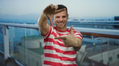 Genç adam çerçeveli bir jest yapıyor, parmakları deniz kenarında bir teknede, çizgili bir gömlek giyiyor, kameraya gülümsüyor, manzaralı bir arka planda gezintiden keyif alıyor..