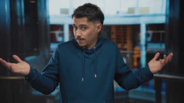 Kapüşonlu genç adam garip bir ifadeyle evde omuz silkiyor, otel ortamında duruyor..