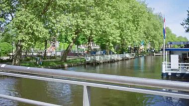 Güneşli bir günde yemyeşil ağaçlar ve teknelerle Amsterdam Kanalı 'nın bulanık görüntüsü Hollanda kentsel manzarasının huzurlu özünü yakalıyor..