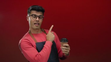 Gözlüklü ve önlüklü genç adam kırmızı arka planda kahve tutuyor, coşku ve güven ifadesiyle işaret ediyor..