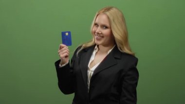 Yeşil arka planda mavi kredi kartı tutan gülümseyen bir kadın siyah takım elbise giyiyor ve finans ve başarı vurguluyor.