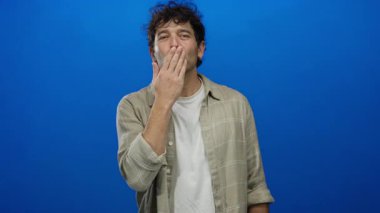 Hispanik bir adam büyüleyici bir gülümseme ile öpücük üflüyor canlı mavi bir arka plana karşı, günlük bej gömlek giyiyor sevgisini ifade ediyor.