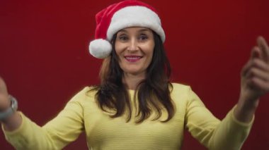 Sarı kazaklı ve Noel Baba şapkalı kadın, kırmızı duvara gülümsüyor, aşağıya bakıyor, neşeli ve neşeli, Noel temalı, izole edilmiş bir arka plan..