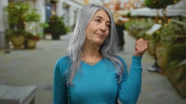 Canlı bir şehir caddesinde gri saçlı yaşlı bir kadın çevrende bitkiler ve insanlarla canlı bir ortamda kendine güvenen bir poz veriyor..