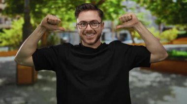 Parkta sakallı ve gözlüklü genç bir adam kendini göstererek enerjik bir şekilde çevresini çevreleyen yeşil ve ağaçlarla çevrili..