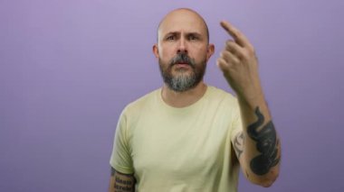 Bald caucasian man with beard gestures with finger against purple background, wearing casual shirt, tattoo visible on arm, isolated setting, serious expression captured.