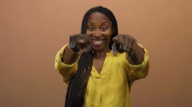 Güvenle gülümseyen kadın, kahverengi arka planda sarı gömlekli kameraya bakıyor. Neşeli bir ifade sergiliyor..