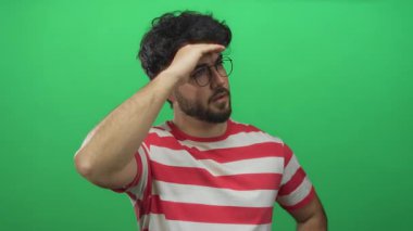 Sakallı ve gözlüklü genç İspanyol adam, çizgili tişört giyen canlı yeşil bir arka planda el ele tutuşup mesafeye bakıyor..