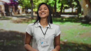 Parkta beyaz gömlekli genç bir kadın yeşil ağaçlar ve parlak bir arka planla açık havada gülümseyerek huzurlu ve neşeli bir ifade sergiliyor..