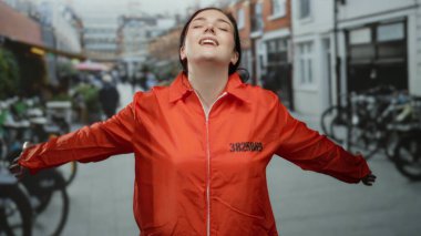 Turuncu hapishane tulumuyla gülümseyen kadın özgürlüğünü kucaklıyor canlı bir şehir caddesinde, neşe ve kurtuluş yayıyor..