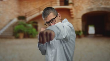 Dışarıda, sokakta dövüşen, gözlüklü, bulanık tuğladan bir binaya bakan bir adam var..