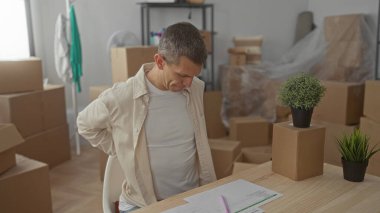 Yeni evinde sırtı ağrıyan bir adam oturma odasındaki kutularla çevrili, masa başında oturmuş, masa başında bitkilerle evrakları gözden geçiriyor, aile içi taşınma anlarını yakalıyor..