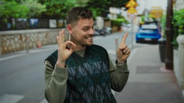 Kenar mahallelerde, yeşilliklerde ve arka planda gün ışığında görülebilen yol işaretleriyle şehir caddesinde gülümseyen bir adam iyi bir jest yapıyor..