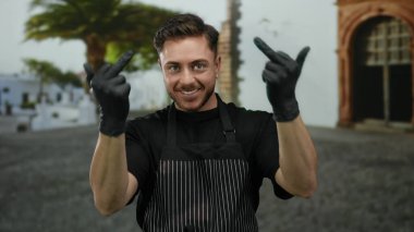 Sakallı genç adam, siyah eldivenler ve çizgili önlükle şehir merkezinde eğlenirken orta parmaklarıyla mizahi hareketler yapıyor..