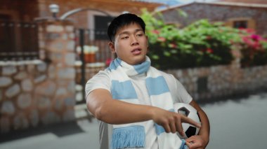 Şehir caddesinde futbol topu tutan genç bir adam uzak bir noktaya doğru hareket ediyor. Canlı şehir ortamında evin yanında eşarp takıyor..