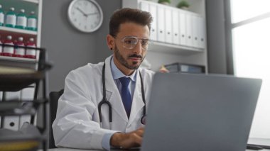 Klinikte çalışan yakışıklı doktor, gözlüklü ve laboratuvar önlüklü, steteskoplu dizüstü bilgisayara odaklanmış, işyerindeki tıp uzmanı..