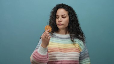 Gökkuşağı süveterli kıvırcık saçlı İspanyol kadın mavi arka planda bir kurabiyeyi inceliyor, izole bir ortamda düşünceli bir yüz ifadesi sergiliyor..