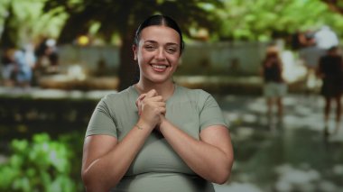 Güneşli bir parkta neşeyle gülümseyen bir kadın yeşil yapraklı ve arka planda yürüyen insanlar, açık havada, kıvrımlı, artı beden ölçülerinde günlük kıyafetli genç bir kadın gösteriyorlar..