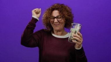 Kahverengi kıvırcık saçlı orta yaşlı bir kadın bir bardak dolusu dolarımızı canlı mor bir arka plana karşı tutarak neşe ve kutlamaları ifade ediyor..