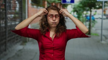 Kırmızı gömlekli kadın kıvırcık saçlı ve gözlüklü duygularını ifade ediyor. Gündüz vakti açık havada sürpriz olduğunu gösteriyor..