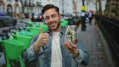 Sakallı ve kot ceketli genç adam şehir caddesinde elinde bir kavanoz dolarla arka planda yeşil scooterlarla baş parmak hareketi yapıyor..