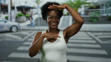 Kadın gülümsüyor ve şehir caddesinde kolsuz gömlekle işaret dili kullanıyor. Arka planda palmiye ağaçları ve arabalar var. Dışarıdaki sakin havayı yansıtıyor..